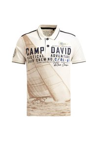 Short-sleeve polo shirt in cream with navy accents. Features a sailboat graphic, text print, and a patch on the left sleeve. Cotton material.
