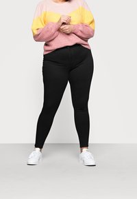 Person med en rosa och gul tröja, svarta tajta byxor och vita sneakers, stående med händerna ihopfattade framför sig mot en vit bakgrund.