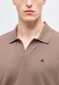 Κοντινό πλάνο ενός ατόμου που φοράει καφέ πουκάμισο με γιακά και μικρό μαύρο λογότυπο «cK» στο αριστερό μέρος του στήθους.