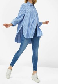Camisa de botones a rayas azul y blanca con mangas largas, que presenta aberturas laterales, combinada con unos vaqueros ajustados azules y zapatillas blancas.