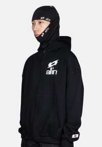 Sweat à fermeture éclair noir en tissu épais, avec une poche avant et un logo sur la poitrine, accompagné d'un masque noir assorti.