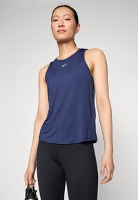 Mornarsko plava bezrukavna atletska majica s okruglim izrezom i bijelim Nike logotipom. Nosiva s crnim tajicama, drži crnu bocu za vodu.