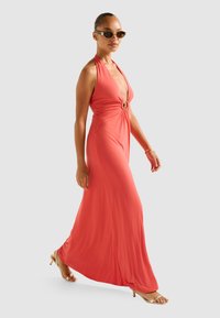 Coral halter maxi dress with a deep V-neck and twist detail at the waist, made of smooth fabric. Paired with gold-heeled sandals.