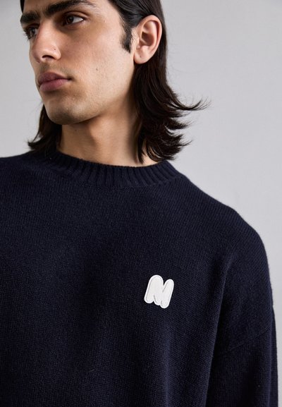 Pulover tricotat bleumarin cu guler rotund și manșete cu nervuri, având un logo brodat alb cu litera "M" pe piept. Textură moale, design casual.