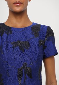 Zila teksturēta kleita ar melniem ziedu rakstiem, īsām piedurknēm un apaļu kakla izgriezumu. Audumam ir savilkta izskata virsma ar spīdumu.