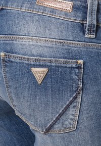 Poche arrière d'un jean en denim bleu avec un logo métallique triangulaire Guess et des détails cousus près du bord de la poche.