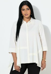 Donna con lunghi capelli neri che indossa orecchini a cerchio dorati, una camicia bianca larga con maniche a 3/4, pantaloni neri e un braccialetto dorato, in piedi contro una parete bianca.