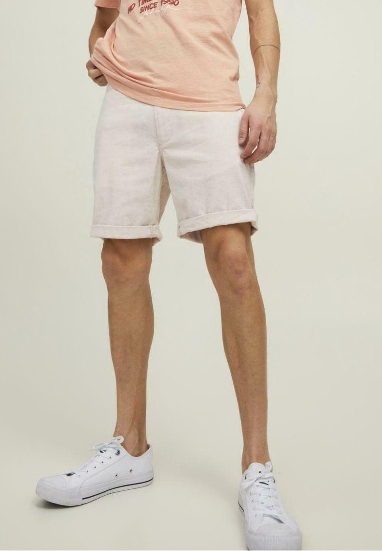 Homme portant un short beige clair retroussé, un t-shirt pêche avec un texte rouge, et des baskets blanches basses, debout devant un fond uni.