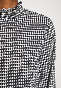 Černobílý bundičkový blůzkový vršek s vysokým límcem a volány na osobě se středně tmavou pletí.