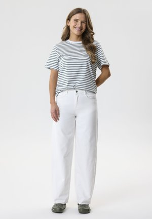 Naeratav naine seisab, kandes valget ja halli triibulist t-särki, valgeid laiade jalgadega pükse ja tumedaid halli sussisid ühevärvilise tausta ees.