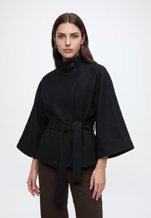Woman with long brown hair wearing a black high-collar jacket with wide sleeves and a tied belt around the waist.
