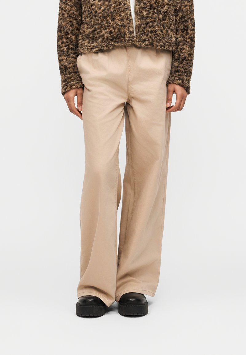 Άτομο που φοράει μπεζ παντελόνι φαρδύ, ένα καφέ cropped σακάκι με υφή και μαύρα χοντρά παπούτσια, στέκεται μπροστά σε απλό φόντο.