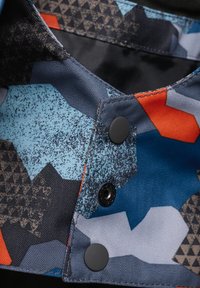 Gemusterter Stoff mit blauen, orangefarbenen und schwarzen geometrischen Designs; verfügt über schwarze Knöpfe und kontrastierende strukturierte Nähdetails.