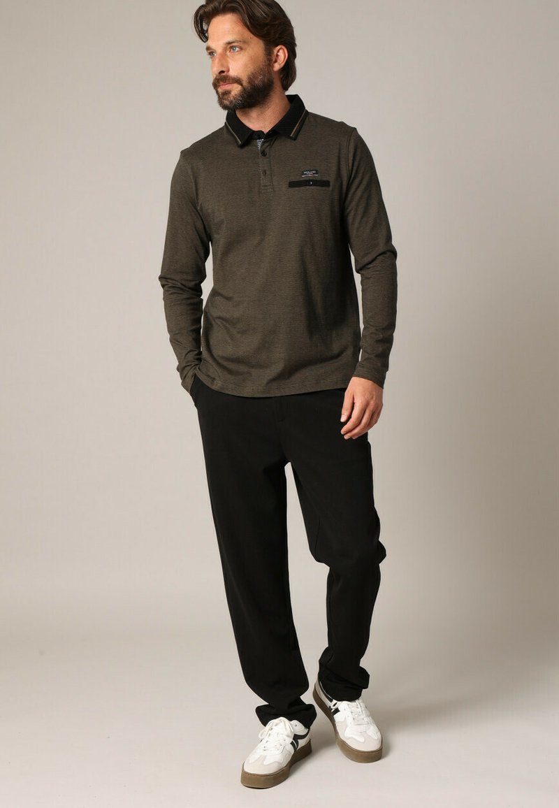 Langärmliges, dunkelolivfarbenes Poloshirt mit schwarzem Kragen und Tasche, kombiniert mit schwarzen Hosen und weißen Sneakers, im lockeren Schnitt.