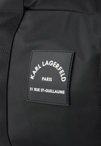 Fekete nejlon táska, amelyen egy téglalap alakú bőrképződmény található fehér szöveggel: "KARL LAGERFELD PARÍZS 21 RUE ST-GUILLAUME" ívelt formátumban.