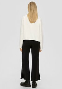 Vrouw met steil blond haar, gekleed in een witte blouse met lange mouwen, zwarte brede broek met zijsplitten en zwarte plateau laarzen, staand met haar rug naar de kijker.