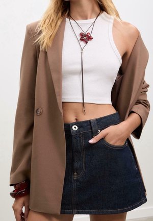 Femme portant un blazer marron sur un crop top blanc, une mini-jupe en jean foncé, un collier de fleurs rouges, et des bracelets rouges à son poignet.