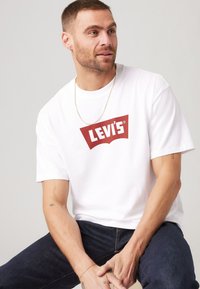 Bela bombažna majica s rdečim logotipom Levi's na prsih, v kombinaciji s temnim jeansom. Preprost dizajn z okroglo ovratnikom in kratkimi rokavi.