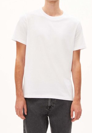 Uomo con t-shirt bianca semplice a maniche corte e jeans grigio scuro, in piedi davanti a uno sfondo chiaro uniforme.