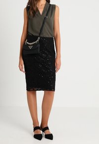 Oliivinvihreä hihaton pusero, yhdistetty mustaan pitsiseen kynähameeseen, jossa on paljettikoristeita. Musta käsilaukku ketjuhihnalla ja teräväkärkiset korkokengät.