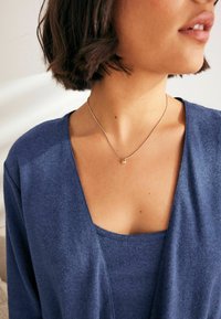 Mėlynas v-neck megztinis, dėvimas ant mėlynos liemenės, papuoštas subtilia sidabrine grandinėle su mažu apvaliu pakabuku. Matomas švelnus tekstūras.