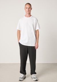 Weißes Baumwoll-T-Shirt mit lockerem Schnitt, kombiniert mit dunkelgrauen Jogginghosen. Das Model trägt graue Turnschuhe. Schlichter Hintergrund.