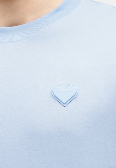 Svetlo modra srajca z majhnim vezenim srčkom na prsih, kjer je izbočen napis blagovne znamke "SANDRO."