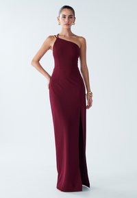 Večerna obleka bordo barve, oprijet kroj, enoramni dizajn, stranski razporek, gladek material, minimalistični detajli, nošena z zlatimi dodatki.