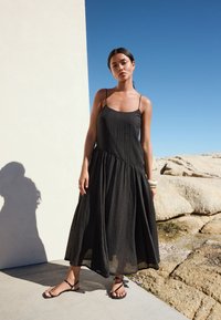 Robe maxi noire sans manches en tissu texturé, silhouette trapèze et ourlet à volants. Portée avec des sandales à brides.