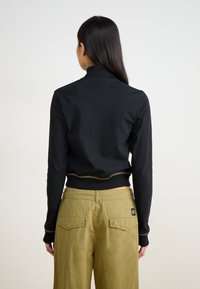 Μαύρη μπλούζα με μακριά μανίκια και ψηλό λαιμό, με στενό σχεδιασμό και αντίθετη ραφή. Συνδυασμένη με ελιά-πράσινο παντελόνι φαρδύ.