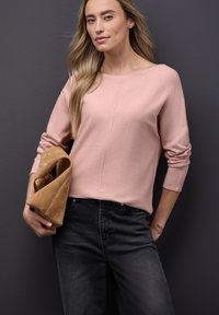 Suéter de manga larga en rosa claro con escote barco, combinado con jeans de mezclilla oscura. La modelo sostiene un clutch de ante color beige plegado.