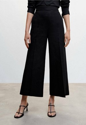 Mujer con pantalones negros de pierna ancha y cortos, y sandalias negras con tiras y tacón, de pie sobre un suelo liso.
