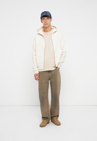 Off-white zip-up hoodie with a tan shirt, olive green pants, and brown sneakers. Denim cap completes the casual outfit.