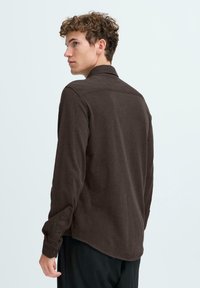 Brun knappestrikket skjorte med krage, lange ermer og buet kant. Mykt stoff med en teksturert finish. Bakvinduet viser detaljer om skulderlær.