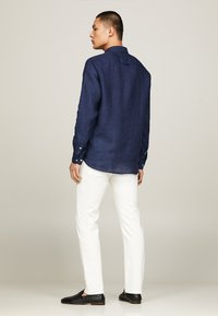 Camicia di lino blu navy con maniche lunghe, polsini a bottoni e orlo arrotondato, abbinata a pantaloni bianchi e scarpe nere.