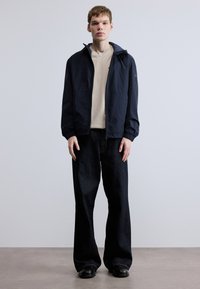 Ľahká námornícka bunda s kapucňou, pod ňou béžové tričko, doplnené širokými tmavomodrými džínsami a čiernymi topánkami. Hladká textúra látky.