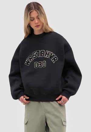Jonge vrouw draagt een zwart oversized sweatshirt met de tekst "WRSTBHVR 030" en lichtgekleurde cargobroek tegen een grijze achtergrond.