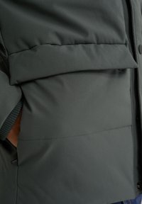 Närbild av en grön vadderad jacka som visar en ficka och strukturerad tyg, vilket framhäver värme och en avslappnad stil.