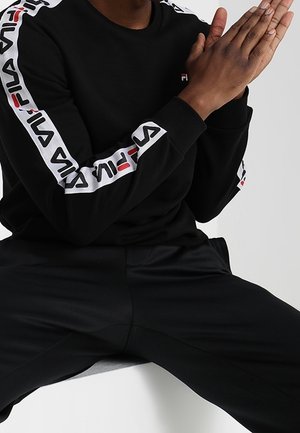 Osoba ubrana w czarny dres FILA z białymi paskami z logo na rękawach, dłonie złączone, siedząca na jednolitym tle.