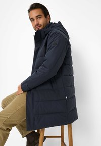 Cappotto lungo navy in piuma con cappuccio, dotato di zip, imbottitura orizzontale e una toppa nera con logo sulla spalla. Abbinato a pantaloni khaki.