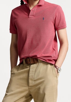 Czerwona koszulka polo z teksturowanej tkaniny, kołnierzykiem na dwa guziki i małym niebieskim logo na piersi. Połączona z beżowymi spodniami i brązowym paskiem.