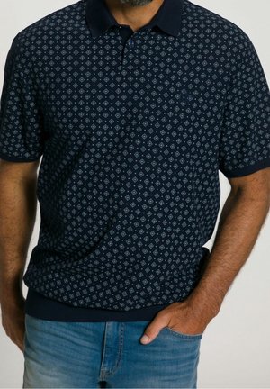 Homme portant un polo à manches courtes bleu marine à motifs avec un col et un jean bleu clair, main glissée dans une poche.