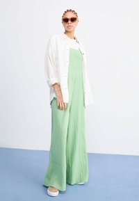 Groene gestructureerde jumpsuit met wijde pijpen en een aansluitend lijfje, gelaagd met een wit overhemd met knopen. Accessoires zijn onder andere bruine zonnebril en tan kleurige schoenen.