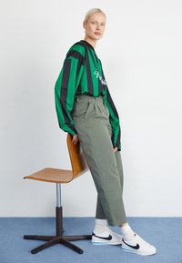 Camicia a maniche lunghe a righe verdi e nere con testo, pantaloni verdi a vita alta e sneakers bianche Nike con dettagli neri, seduto su una sedia di legno.