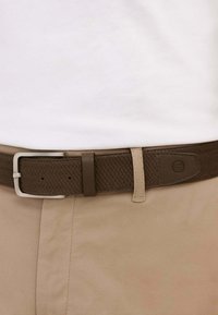 Cintura intrecciata in pelle marrone con fibbia in metallo argentato; presenta una texture liscia e un dettaglio di cucitura sottile, abbinata a pantaloni beige.