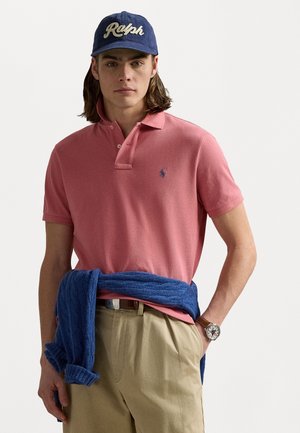 Rozā kokvilnas polo krekls ar īsām piedurknēm un zilu polo logotipu, kombinēts ar bēša biksēm un zilu adītu džemperi, kas saistīts ap jostasvietu.