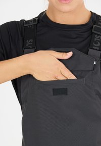 Schwarze Latzhose mit verstellbaren Trägern, ausgestattet mit einer Vorderseite-Tasche mit einem Klettverschluss-Deckel. Das Material wirkt glatt und langlebig.