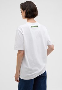 T-shirt branco de algodão com gola redonda, mangas curtas e um rótulo verde na nuca. O tecido é suave e sem textura.