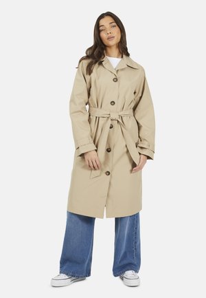 Femme portant un trench-coat beige ceinturé, un jean bleu à jambes larges et des baskets blanches, debout devant un fond clair uni.
