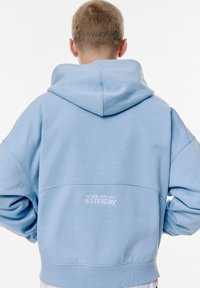 Haut à capuche bleu clair en tissu doux, doté d'une grande capuche, d'épaules tombantes et d'un texte brodé au dos indiquant "L'AVENIR EST ARRIVÉ HIER."
